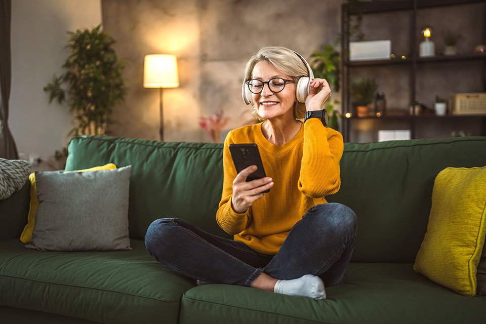 Eine reife blonde Frau hört mit Kopfhörern Musik auf dem Sofa (Foto: Miljan Zivkovic/Shutterstock.com) 