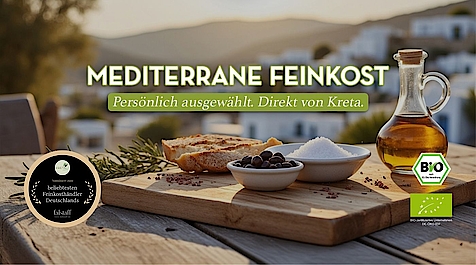 gedeckter Holztisch mit Brot, Oliven, Salz und Olivenöl vor mediterraner Landschaft, Text „Natürlich, nachhaltig & fair – Mediterrane Feinkost“ und Bio-Siegel u. Feinkost-Auszeichnung.