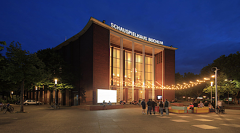 Beleuchtetes Schauspielhaus Bochum bei Nacht, mit Menschen und Lichterketten davor.