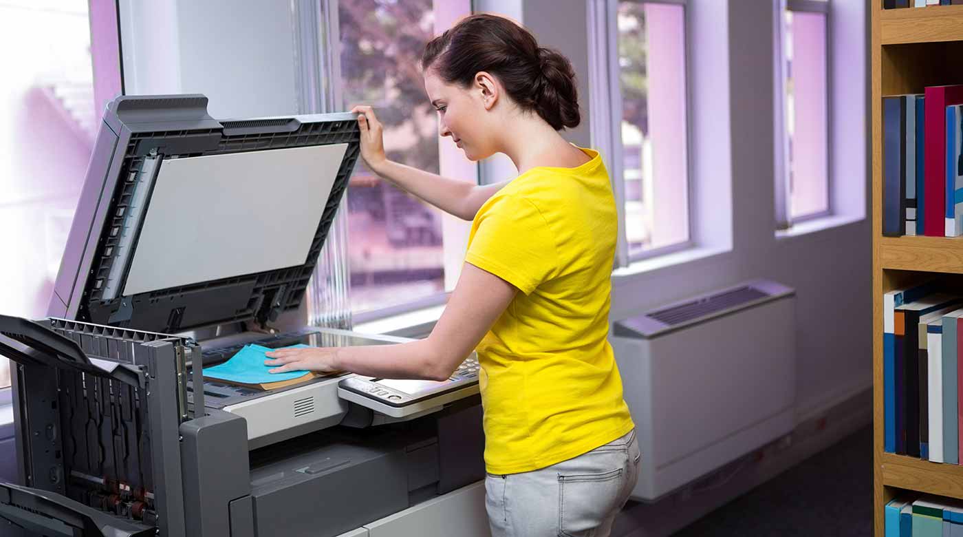Frau in gelbem Shirt bedient einen Kopierer in einem hellen Raum mit Bücherregal.