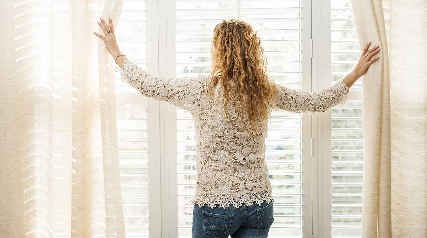 Frau öffnet Vorhänge vor stilvollem Fenster mit Lamellen, betont elegante Raumgestaltung.